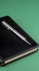 A sleek silver ballpoint pen resting on a closed black notebook with a green background, showcasing a professional and organized workspace setup