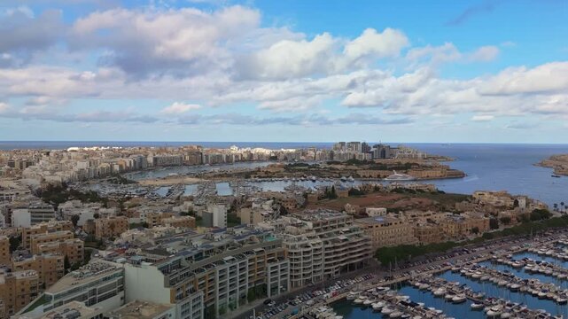 Aerial view over Msida Marina and Ta&rsquo; Xbiex, Malta, looking toward Sliema and Manoel Island with yachts, coastal skyline, and calm Mediterranean waters under bright partly cloudy skies.