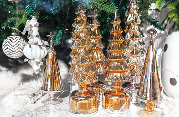 Metallic gold Christmas trees of various shapes sit on a snowy white surface, glowing with string lights. Pine branches and elegant ornaments in background enhance festive, cozy holiday atmosphere