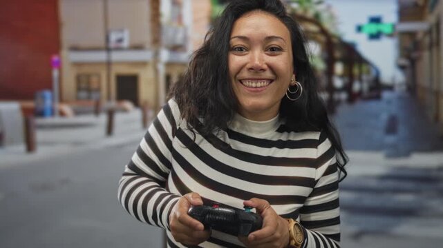 Woman holding a game controller with both hands, thumbs pressing buttons, standing on a city street near shopfronts; playful joy.