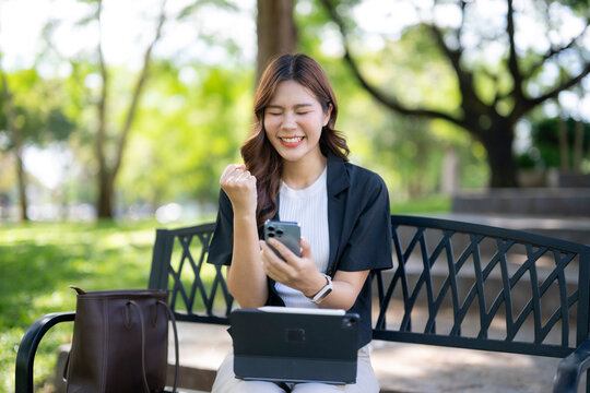 Asian woman celebrating success using phone in park