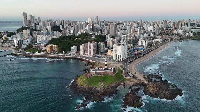 Speed-up aerial drone video of the Farol da Barra lighthouse and the surrounding coastline of Salvador, Bahia, Brazil. The footage includes a wide city view with tall modern buildings.