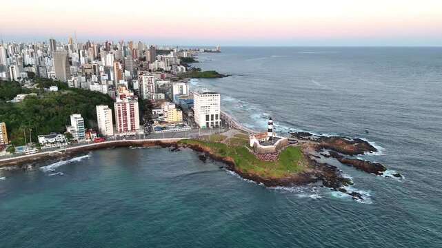 Aerial drone orbit of Farol da Barra lighthouse and the Barra coastline with Salvador city skyline in the background