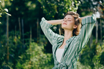 Woman outdoors, eyes closed, basking in sunlight with a striped green shirt, standing in a lush...