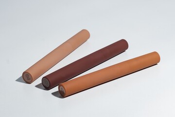 Three matte nude tone cosmetic tubes, possibly mascara or concealer, arranged diagonally on a white background with subtle shadows. Minimalist beauty product display.