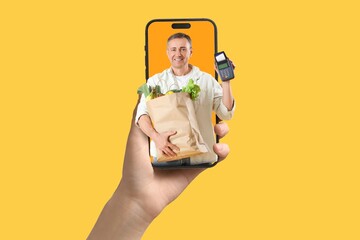 Hand holding mobile phone with male cashier with grocery bag on screen against yellow background