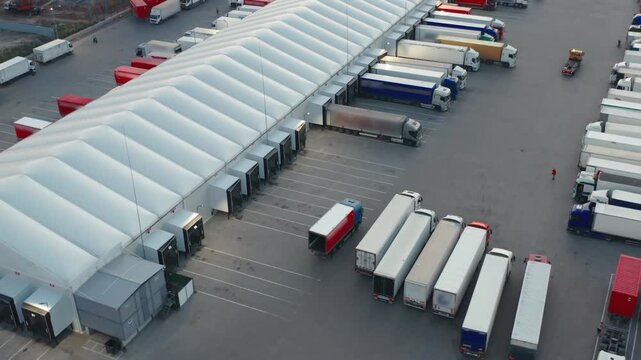 Aerial view of a modern logistics warehouse with multiple semi-trailers trucks parked at ramps for loading and unloading goods