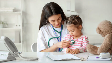 Female pediatrician with stethoscope listening to cute little girl at table in clinic