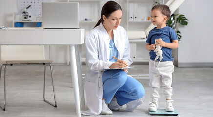 Female pediatrician with cute little boy measuring weight on scales in clinic
