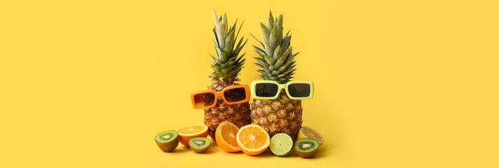 Fototapeta premium Fresh pineapples with sunglasses, different citrus fruits and kiwi on yellow background