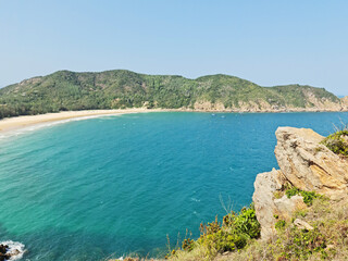 A breathtaking view of a crescent-shaped beach and the vibrant blue-green ocean, framed by a large foreground rock and forested hills