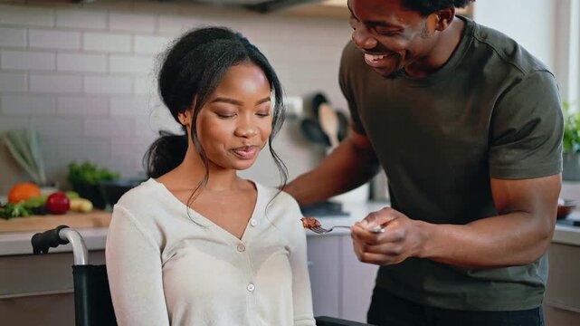 Caring black man feeding his smiling partner in a wheelchair in the kitchen, letting her taste the food while preparing a meal together, she nods with her head as a signal of approval