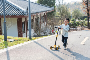 A young boy is enjoying riding a scooter on a paved path in an outdoor park with a traditional building in the background.