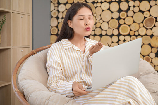 Young Asian woman with laptop having online date in armchair at home