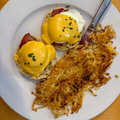 Classic Eggs Benedict Breakfast. Rich Hollandaise Sauce and Poached Eggs Served with Crispy, Well-Done Hash Browns on a White Plate, Overhead View