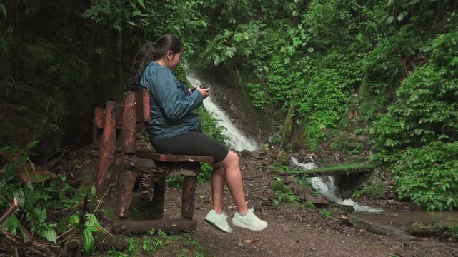 A woman sits on a rustic wooden bench, calmly reviewing the photos on her camera while a beautiful waterfall flows in the background, creating a peaceful outdoor moment.