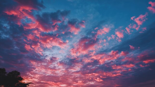 A vibrant sunset with pink and blue hues, featuring a dramatic sky with clouds and a silhouette of trees in the foreground.