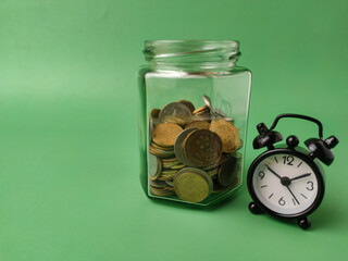 A retro style alarm clock in front of a glass jar filled with coins