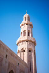 A striking architectural photograph of the minaret tower at the Sultan Qaboos Grand Mosque in Muscat, Oman. The image highlights traditional Islamic design elements, including arched openings