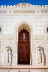 A striking architectural photograph showcasing an ornate wooden entrance door framed by an intricately carved marble archway at the Sultan Qaboos Grand Mosque in Muscat, Oman