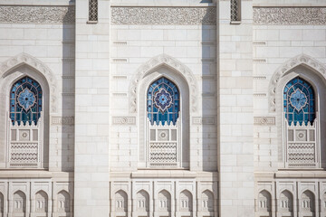 A beautifully symmetrical architectural photograph featuring three ornate stained-glass windows framed by intricate Islamic carvings and white marble walls at the Sultan Qaboos Grand Mosque in Muscat