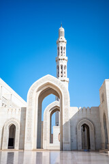 A bright and elegant photograph showcasing the towering minaret and ornate arched gateways of the Sultan Qaboos Grand Mosque in Muscat, Oman. The white marble architecture, detailed Islamic carvings