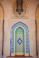 A vibrant close-up photograph of an intricately designed Islamic mosaic tile niche at the Sultan Qaboos Grand Mosque in Muscat, Oman. The composition features vivid blue, turquoise, and gold geometric
