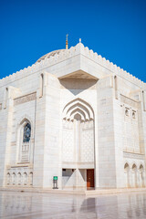 A stunning close-up photograph of the white marble exterior of the Sultan Qaboos Grand Mosque in Muscat, Oman. The image captures intricate Islamic architecture, geometric carvings, arched doorways