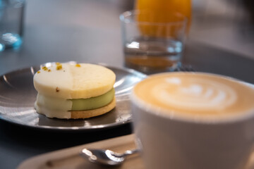 Close-up of a horizontal photo of a barista cafe focusing on a pistachio alfajor with white chocolate and orange juice in the background