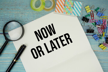 Now Or Later text on a paper surrounded by office supplies and magnifying glass on a blue wooden...