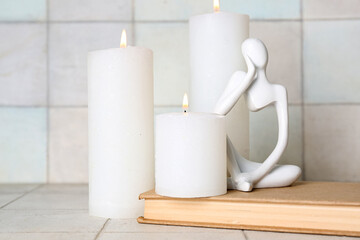 Burning candles with decorative human figure and book on white tile background