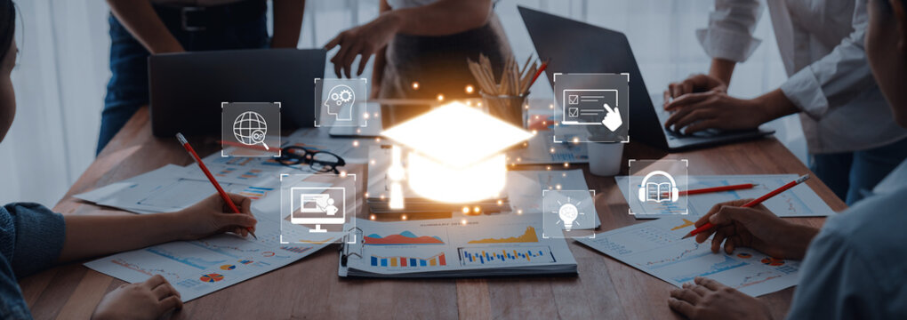 A dynamic team collaborates around a desk filled with laptops and papers, surrounded by educational icons, showcasing innovative approaches to learning in a professional environment. Trope - Powered by Adobe