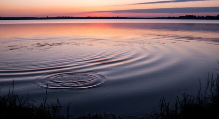A serene lakeside scene at sunset with calm water reflecting the colorful sky and gentle ripples near the shoreline