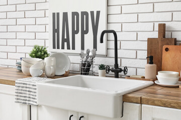 Wooden kitchen counter with ceramic sink, clean dishes and poster hanging on light brick wall