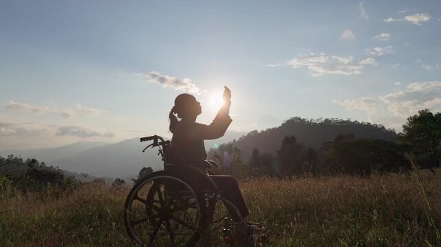 Silhouette Disabled handicapped woman sitting on wheelchair praying to God on mountain sunset background.