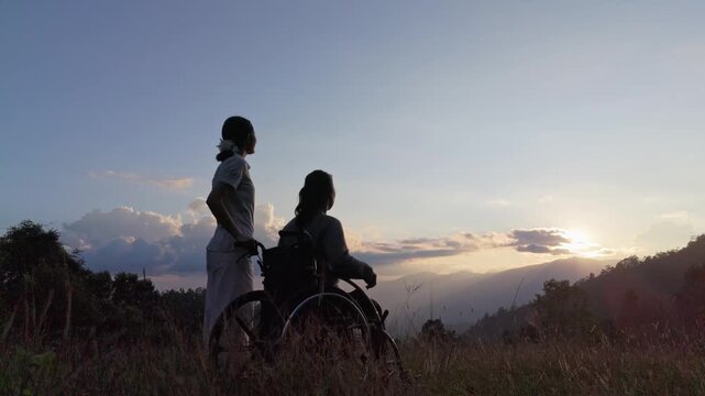 Silhouette of elderly woman in wheelchair with her care helper looking at sunset mountain