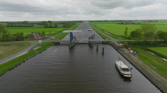 Approach view toward a bascule (draw) bridge on a straight Dutch canal, with farmland, service road and small craft visible under grey skies.