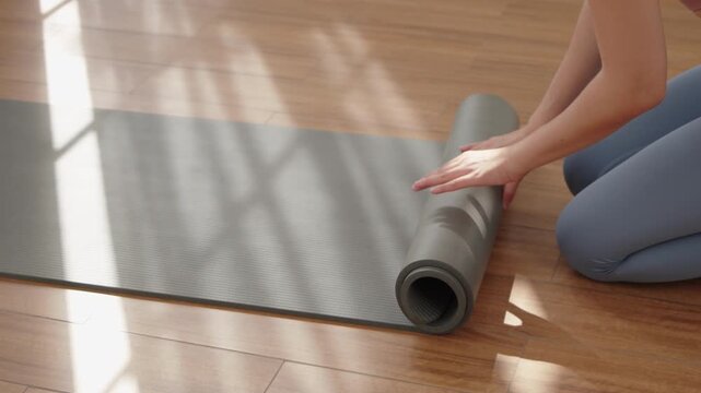 Close up Asian woman hands rolling her yoga mat after workout as the morning light the wooden floor, showing a simple asian moment of calm and routine with exercising.