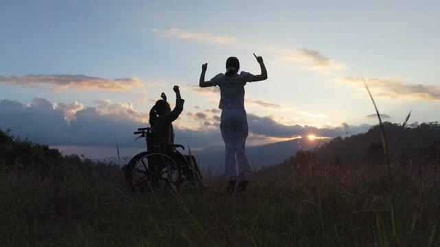 Silhouette elderly woman in wheelchair dancing with his care helper on mountain sunset. Psychological medication  treatment concept