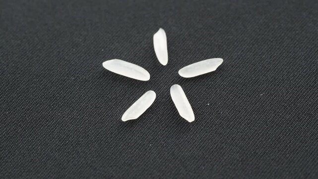 Top view 4K loop of five ST25 rice grains arranged in a star/flower pattern. The scene rotates slowly on a minimal black background. Conceptual video for premium food and design.