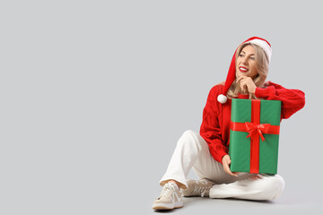 Beautiful middle-aged woman with Christmas gift sitting on grey background