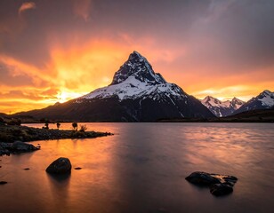 A majestic snow-capped mountain reflects in calm water under a vibrant orange and golden sunset sky. Some rocks dot the foreground