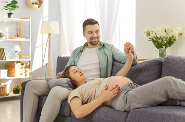 Happy young couple relaxing on sofa at home with pregnant woman lying on lap of her husband and touching belly. Smiling future parents holding hands and sharing tender moment while awaiting baby.
