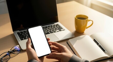 Person holds smartphone with blank screen next to laptop and coffee
