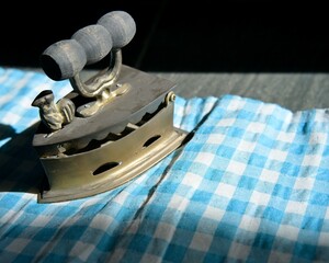 An antique brass iron exposed to sunlight on blue patterned fabric. An old household ironing tool.