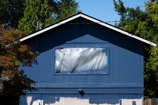 Residential house being painted dark blue, window covered and protected by plastic, sunny summer day in neighborhood
