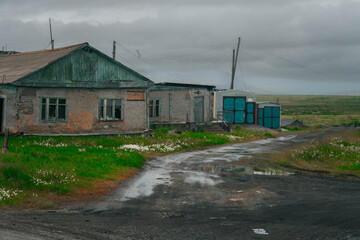 Abandoned wooden house and garages in Ugolnye Kopi, Chukotka, Russia, beside muddy road and tundra fields of wildflowers.