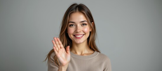 Smiling woman waving hello, wearing a cozy sweater against a neutral background.