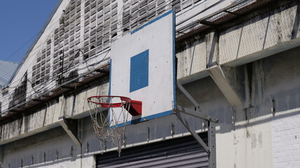 Outdoor basketball hoop and backboard mounted on the textured, weathered facade of an industrial warehouse building under a blue sky