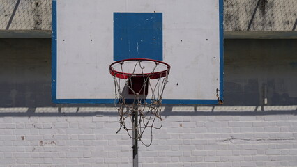 Weathered outdoor basketball hoop, featuring a red rim and worn net set against a white and blue backboard above a sunlit, white-painted brick wall